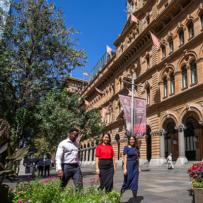 Three people walking on a sunny day in front of a historic building with arched windows and flags. Vibrant greenery and colorful flowers line the sidewalk.