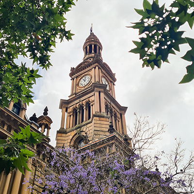 Historic clock tower with a copper roof stands tall, framed by green leaves and purple-bloomed branches against a cloudy sky.